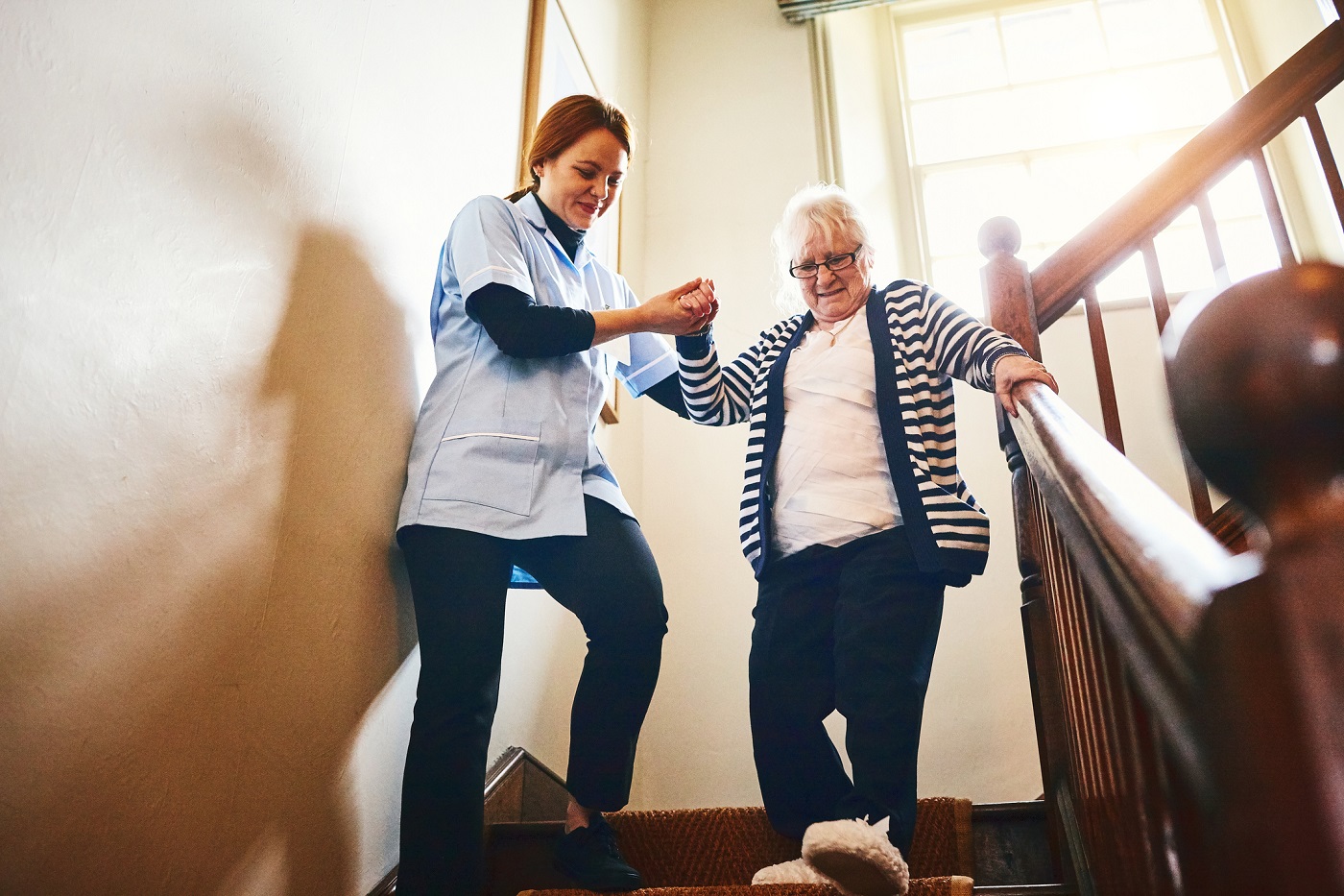 Young female caregiver helping senior woman walking down stairs at home.
